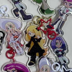 Puede incluir: Colección de llaveros acrílicos transparentes con varios personajes de anime. Los llaveros tienen diseños coloridos, incluyendo personajes con pelo morado, rubio y verde. Algunos llaveros tienen detalles rojos en forma de estrella y cierres metálicos.