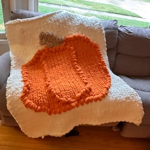 May include: A chunky knit throw blanket featuring a large, textured orange pumpkin design centred on a solid white background. The pumpkin has a small, light brown stem at the top, and the entire piece is crafted from thick, soft yarn loops.