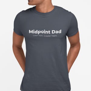 Puede incluir: Una camiseta gris oscuro con el texto blanco "Midpoint Dad" y la frase "Later start, Greater heart." La camiseta es de cuello redondo y parece estar hecha de un material suave. El diseño es simple y moderno.