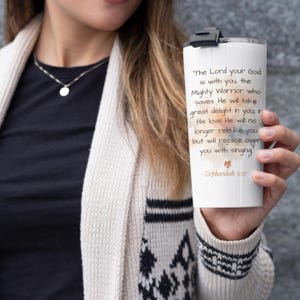 May include: White travel tumbler with a black lid, featuring the text "The Lord your God is with you..." from Zephaniah 3:17. The tumbler has a black closure and is held by a person wearing a cream-colored cardigan.