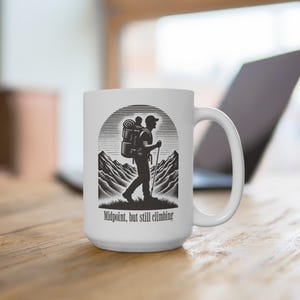 Puede incluir: Taza de cerámica blanca con una ilustración en blanco y negro de un excursionista con mochila y bastón, sobre un fondo de montañas. El texto "Midpoint, but still climbing" está impreso debajo. La taza tiene un asa grande.