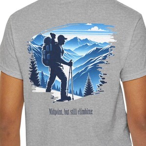 Puede incluir: Camiseta gris con un diseño de montaña azul y blanco. El diseño muestra a un excursionista con una mochila y bastones de senderismo. El texto "Midpoint, but still climbing" está impreso debajo.