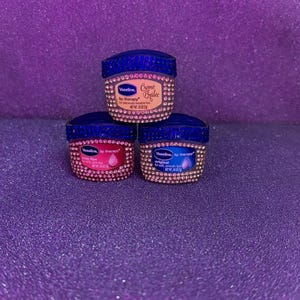 May include: Three Vaseline Lip Therapy containers decorated with blue and clear rhinestones. The top container is Crème Brûlée, the bottom left is Rosy Lips, and the bottom right is Original. Each container has a net weight of 0.25 ounces.