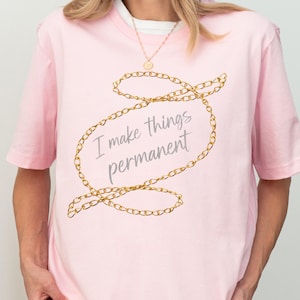 May include: A pale pink t-shirt featuring the phrase "I make things permanent" in a gold chain design. The shirt is a soft, light pink colour. The words "Soft- Light Pink" are written in cursive at the bottom left.