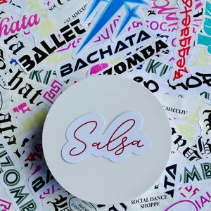 May include: A white round sticker with the word "Salsa" written in red cursive lettering. The sticker is surrounded by other colorful stickers with words like "Bachata", "Ballet", and "Zomba" written on them.