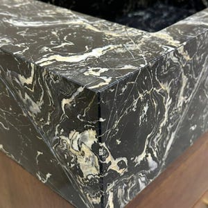 May include: A black marble sink basin featuring prominent white and cream-colored veining throughout the stone surface. The basin has sharp, clean edges and a polished finish, mounted atop a wooden cabinet base with a visible grain pattern.