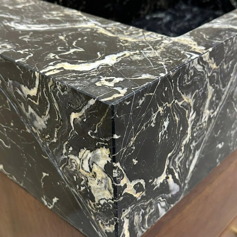 May include: A black marble sink basin featuring prominent white and cream-colored veining throughout the stone surface. The basin has sharp, clean edges and a polished finish, mounted atop a wooden cabinet base with a visible grain pattern.