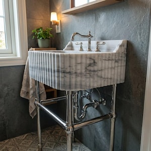 May include: A white marble pedestal sink with a fluted exterior design and gray veining. The sink features a polished chrome faucet with two matching handles and is supported by a chrome metal frame with exposed plumbing pipes underneath.