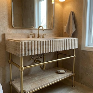 Op de afbeelding: Een badkamermeubel met een beige, gecanneleerde stenen wastafel, ondersteund door een frame van geborstelde goudkleurige metalen buizen. Het meubel bevat een onderste stenen plank, een bijpassende gouden kraan en een rechthoekige spiegel erboven.