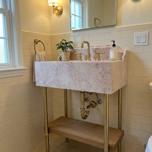 May include: A pink marble bathroom sink with a brass tap and plumbing fixtures. The sink is supported by a brass metal frame with a wooden shelf underneath. A small potted plant sits on the back ledge of the sink next to a soap dispenser.