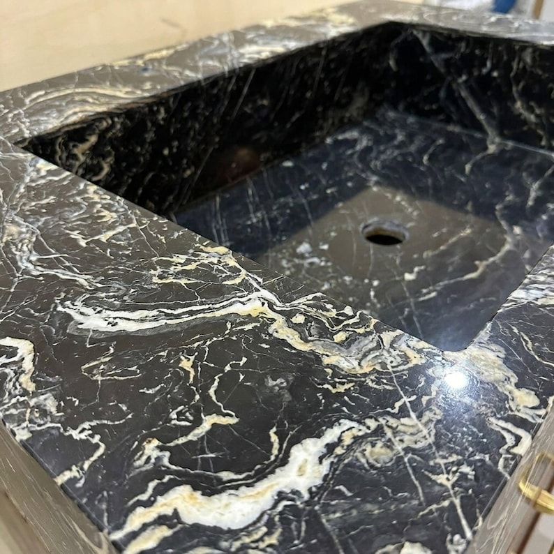 May include: A rectangular bathroom sink crafted from polished black marble featuring prominent white and gold veining throughout the stone surface. The basin includes a centered circular drain hole and thick, smooth edges with a natural stone pattern.