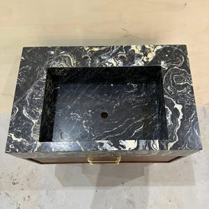 May include: A rectangular bathroom vanity featuring a black marble countertop with prominent white and gold veining. The basin is integrated into the stone surface, which includes two faucet holes. A wooden cabinet base sits below with a gold handle.