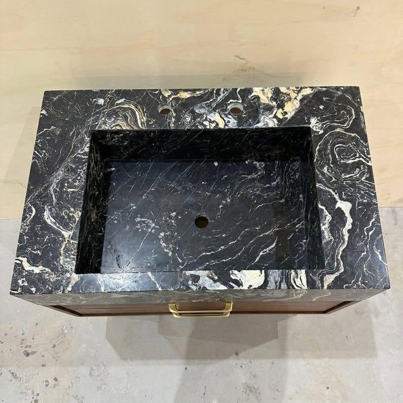 May include: A rectangular bathroom vanity featuring a black marble countertop with prominent white and gold veining. The basin is integrated into the stone surface, which includes two faucet holes. A wooden cabinet base sits below with a gold handle.