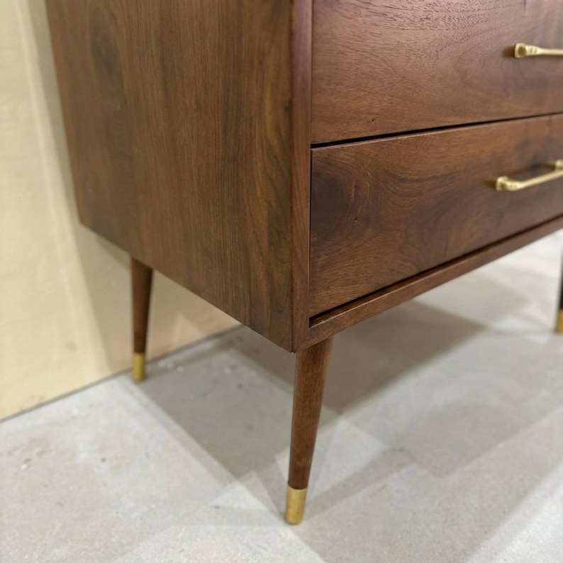 May include: A dark brown wooden chest of drawers featuring two horizontal drawers with thin, horizontal gold-toned metal handles. The piece is supported by four tapered wooden legs, each finished with a gold-toned metal cap at the bottom.