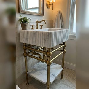 May include: A white marble sink with a fluted exterior design, supported by a brass metal frame with a lower marble shelf. The sink features a brass faucet and two matching handles, with a small potted green plant placed on the corner.
