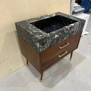 May include: A bathroom vanity featuring a dark brown wooden cabinet with two drawers, gold-toned horizontal handles, and four tapered legs with gold-toned feet. The top is a thick, rectangular black marble basin with prominent white veining.