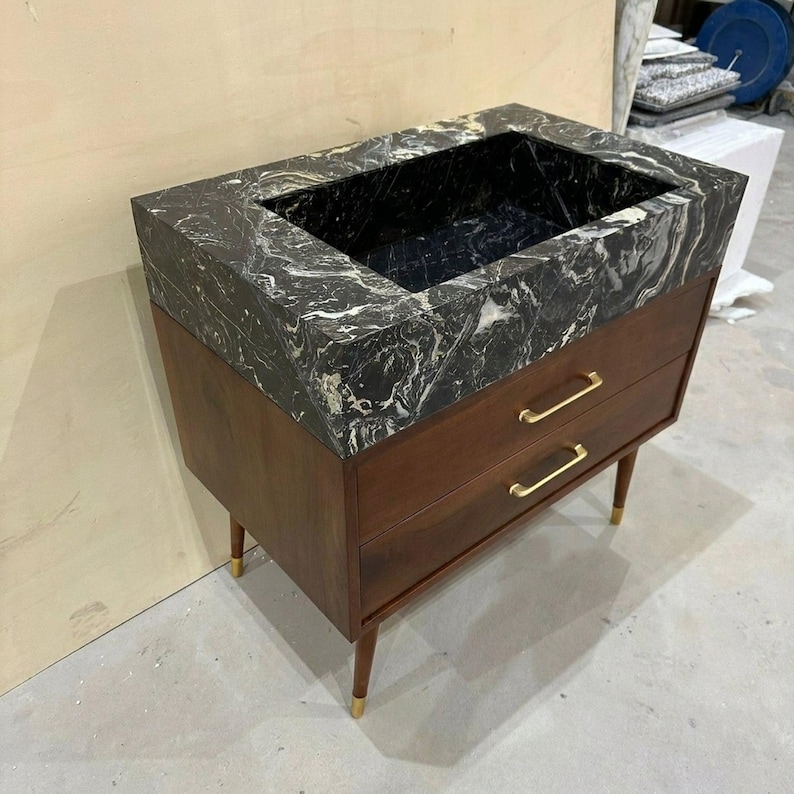 May include: A bathroom vanity featuring a dark brown wooden cabinet with two drawers, gold-toned horizontal handles, and four tapered legs with gold-toned feet. The top is a thick, rectangular black marble basin with prominent white veining.