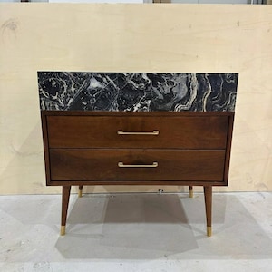 May include: A wooden chest of drawers featuring two horizontal drawers with thin gold-toned metal handles. The top surface is a slab of black and white veined marble. The unit is supported by four tapered wooden legs with gold-toned metal feet.