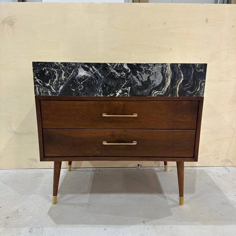May include: A wooden chest of drawers featuring two horizontal drawers with thin gold-toned metal handles. The top surface is a slab of black and white veined marble. The unit is supported by four tapered wooden legs with gold-toned metal feet.