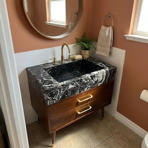 May include: A bathroom vanity featuring a dark wood cabinet with two drawers, gold-toned handles, and tapered legs. The countertop is black marble with white veining, paired with a gold faucet, a small potted green plant, and a bar of soap.