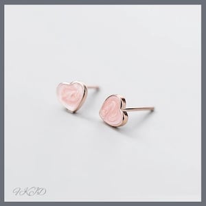 Peut inclure: Une paire de boucles d'oreilles en forme de cœur. Les boucles d'oreilles ont une finition en or rose avec un remplissage en émail rose clair. Les boucles d'oreilles sont présentées sur un fond gris clair. La marque "FKID" est visible en bas à gauche.