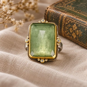 May include: A ring featuring a large, rectangular, faceted light green stone set in a gold-toned bezel. The setting is accented with small, clear round stones arranged in clusters at the top, bottom, and sides, mounted on a dark metal band.