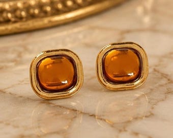 May include: A pair of stud earrings featuring square-shaped amber-colored glass stones set within polished gold-tone metal frames. The earrings have rounded corners and a smooth, reflective surface, showcasing a warm, translucent orange hue.