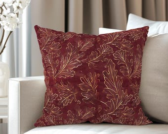 May include: A square decorative pillow with a burgundy background and a gold leaf pattern. The pillow is on a white sofa. A white vase with white flowers is on a side table.