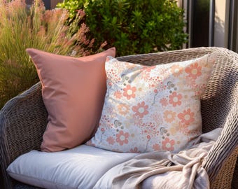 Pode incluir: Duas almofadas decorativas numa cadeira de vime. Uma almofada é pêssego liso, a outra apresenta um padrão floral em pêssego, branco e cinza. Uma manta bege está sobre a cadeira.