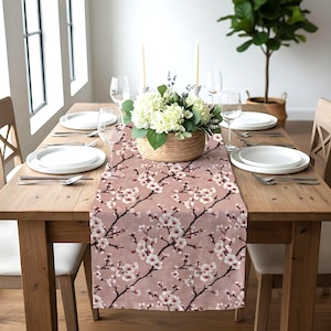 May include: A floral table runner with a pink background and a pattern of white cherry blossoms. The runner is placed on a wooden table set for a meal, with white plates, silverware, and wine glasses. A basket of flowers sits in the center.