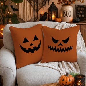 May include: Two square orange throw pillows featuring black jack-o'-lantern face designs. One pillow displays a smiling face with triangular eyes, while the other features a jagged, menacing grin with sharp, angular eyes printed on the fabric.