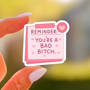 Puede incluir: Una pegatina rosa con forma de nota adhesiva con el texto "REMINDER: YOU'RE A BAD BITCH." escrito a mano. La pegatina tiene un gráfico de corazón rosa y pequeños corazones rosas esparcidos alrededor del texto.