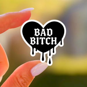 May include: A black heart-shaped sticker with the words "BAD BITCH" in white, dripping at the bottom. The sticker is held between two fingers against a blurred background.