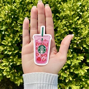 May include: A pink sticker of a Starbucks iced drink with strawberries and a green straw. The Starbucks logo is visible on the cup. The sticker is held in front of a green leafy background.