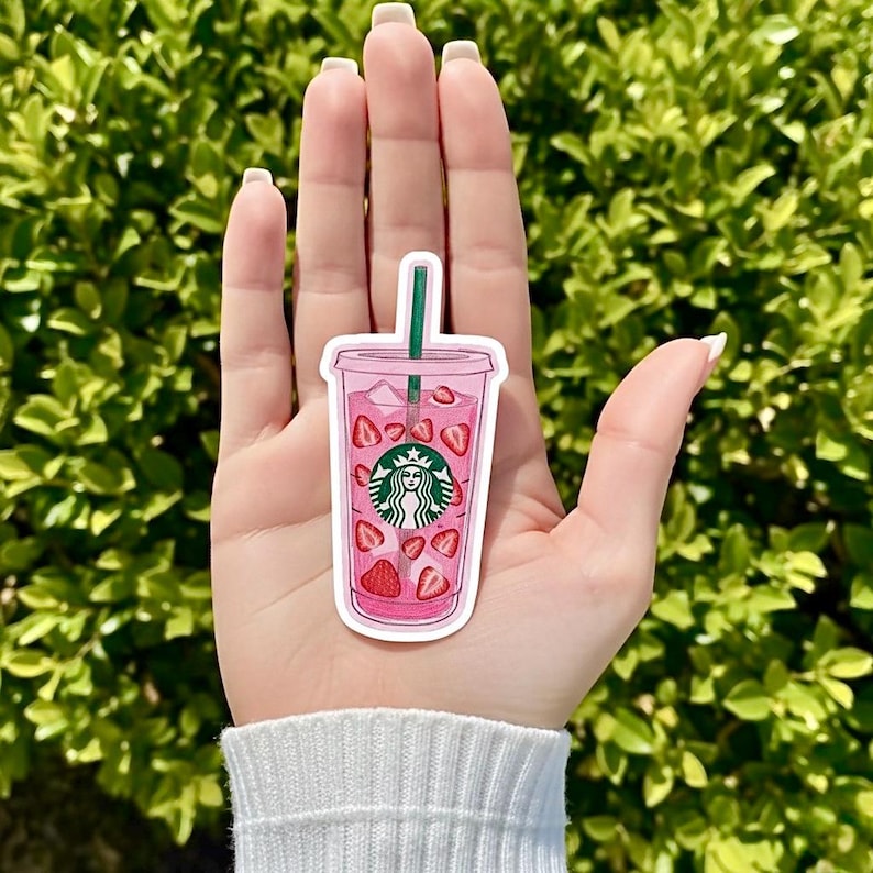 May include: A pink sticker of a Starbucks iced drink with strawberries and a green straw. The Starbucks logo is visible on the cup. The sticker is held in front of a green leafy background.