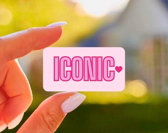 May include: A pink rectangular sticker with the word "ICONIC" in a bold, pink font. A small pink heart is on the right side of the word. The sticker is held between two fingers against a blurred background of green and a house.