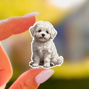 May include: A white, fluffy puppy sticker with a cute expression. The sticker features a sitting dog with a pink tongue sticking out. The dog has dark eyes and a fluffy coat. The sticker is held between two fingers.