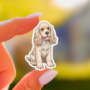 Puede incluir: Una pegatina con una ilustración de dibujos animados de un cachorro de Cocker Spaniel. El perro es de color marrón claro con orejas largas y onduladas y una expresión feliz, con la lengua fuera. La pegatina se sostiene entre dos dedos sobre un fondo amarillo y verde borroso.