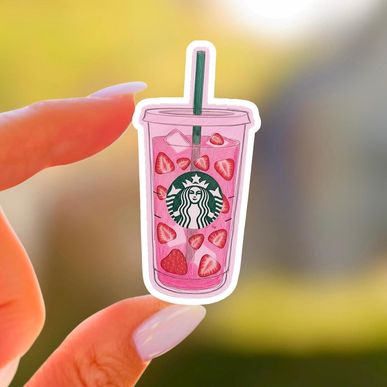 May include: A pink sticker featuring a Starbucks-style cup filled with pink liquid and strawberry slices. The cup has a green straw and the Starbucks logo. The sticker is held between two fingers.