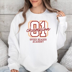 Può includere: Felpa bianca con la scritta "01 champion" in bordeaux e pesca. Sotto, la scritta "SPEED READER ACADEMY" è in bordeaux. La felpa è a girocollo.