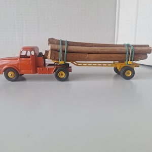 Pode incluir: Um caminhão de brinquedo vintage laranja com um reboque amarelo transportando toras de madeira. O caminhão tem pneus pretos com centros amarelos. As toras são presas com faixas verdes. Um brinquedo clássico.