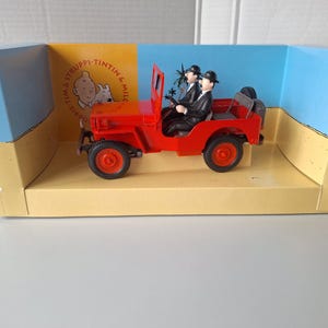 May include: A red toy jeep with two figures in black suits and bowler hats. The jeep is in a display box with the words "Tintin & Snowy" and a cartoon image. The box has a yellow and blue background.