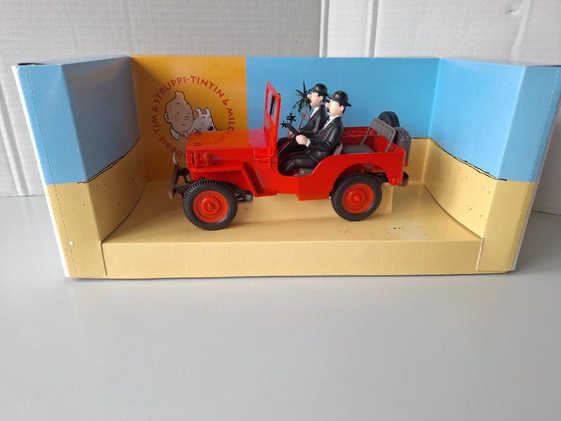 May include: A red toy jeep with two figures in black suits and bowler hats. The jeep is in a display box with the words "Tintin & Snowy" and a cartoon image. The box has a yellow and blue background.