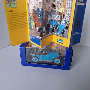 Puede incluir: Un coche de juguete azul de la serie Tint&iacute;n, exhibido en una caja azul con un libro amarillo y azul. El libro presenta ilustraciones de una escena de desfile y texto en holand&eacute;s. El coche es de estilo vintage.