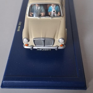 Op de afbeelding: Een beige modelauto met twee figuren erin, &eacute;&eacute;n in een pak en de ander in een blauw overhemd. De auto staat op een donkerblauwe basis. De nummerplaat van de auto luidt HML52511.