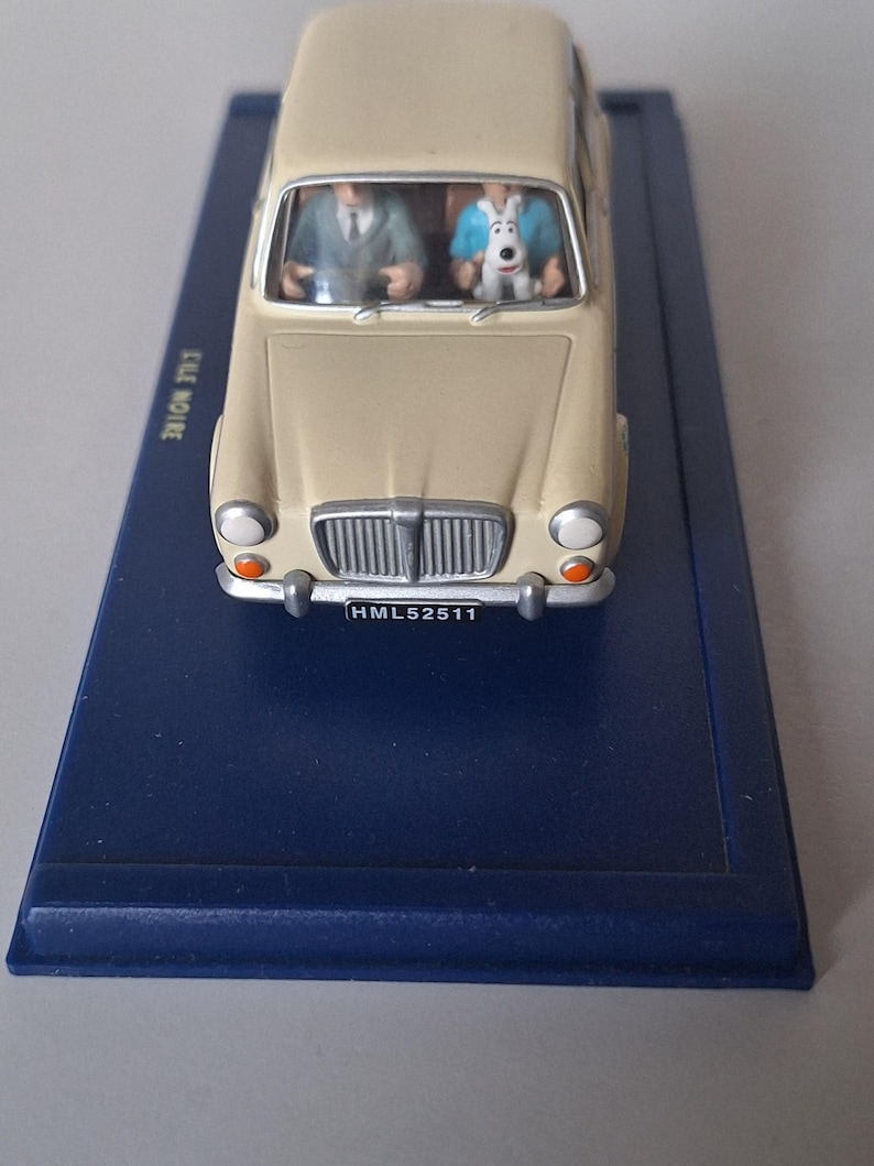 Op de afbeelding: Een beige modelauto met twee figuren erin, &eacute;&eacute;n in een pak en de ander in een blauw overhemd. De auto staat op een donkerblauwe basis. De nummerplaat van de auto luidt HML52511.