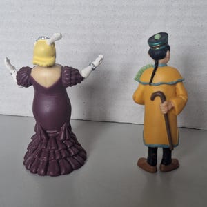 May include: Two figurines, one of a woman in a purple dress with her arms outstretched, and another of a man in a yellow coat holding a cane. The woman has yellow hair and the man has a black hat. The figurines are on a gray surface.