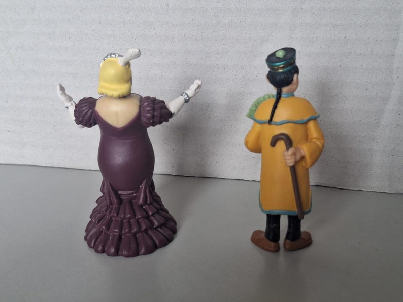 May include: Two figurines, one of a woman in a purple dress with her arms outstretched, and another of a man in a yellow coat holding a cane. The woman has yellow hair and the man has a black hat. The figurines are on a gray surface.