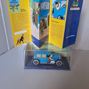 Może przedstawiać: Niebieski model taks&oacute;wki z serii Tintin w Ameryce, z figurką Tintina i Milusia. Model jest prezentowany w przezroczystej obudowie, w towarzystwie złożonego komiksu z ilustracjami i tekstem.