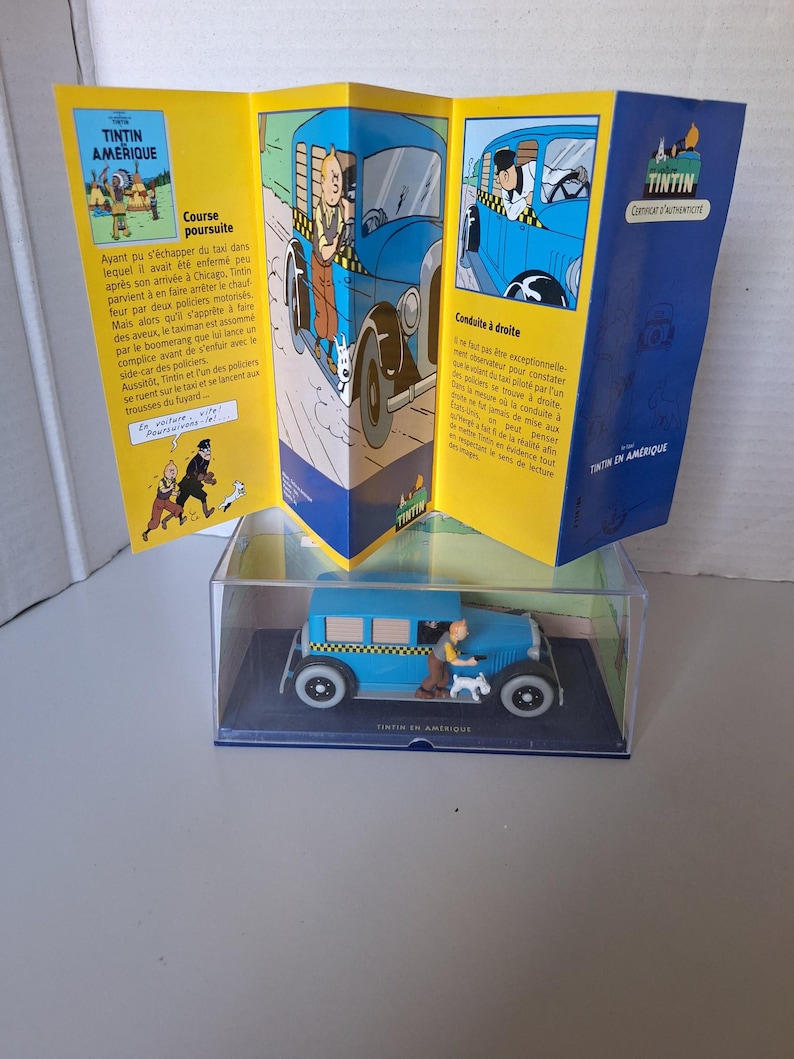 Może przedstawiać: Niebieski model taks&oacute;wki z serii Tintin w Ameryce, z figurką Tintina i Milusia. Model jest prezentowany w przezroczystej obudowie, w towarzystwie złożonego komiksu z ilustracjami i tekstem.
